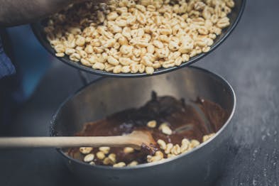 Gepuffter Weizen wird zu Schokomasse in eine Metallschale mit Kochlöffel gegeben