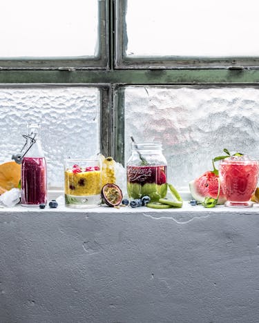 Auf einem weißen Fensterbrett stehen verschiedene Gläser mit Smoothies und Säften. Verziert mit frischen Beeren und Früchten