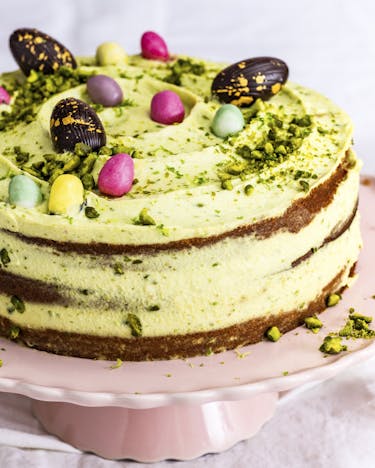 Eine mehrstöckige Torte mit gelb-grüner Creme eingestrichen, garniert mit bunten Ostereiern. Die Torte steht auf einer rosa Tortenplatte mit erhöhtem Fuß.