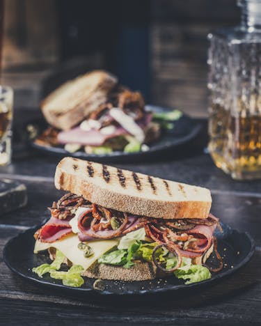 Pastrami Sandwich auf einem schwarzen Teller, im Hintergrund volle Gläser, eine Whiskeyflasche und ein weiterer Teller mit einem Sandwich