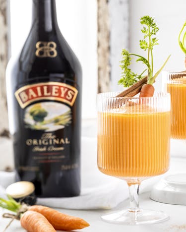 Knall-orangene Smoothies sind in zwei Gläsern serviert und angerichtet mit je einer kleinen Karotte und Zimtstangen. Daneben steht eine Flasche Baileys.