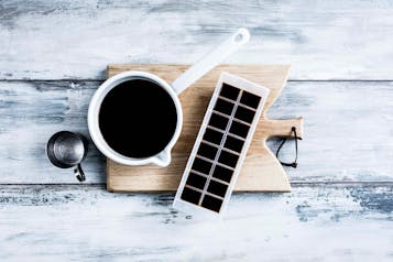Auf einem Holzbrett steht ein Becher mit Kaffee, daneben eine Eiswürfelform mit Kaffee gefüllt