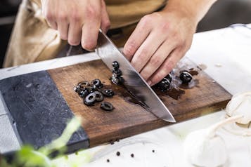 Hände, die auf einem Holzbrett schwarze Oliven in Scheiben schneiden