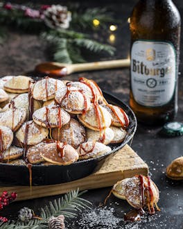 Glutenfreie Poffertjes mit Bier-Karamell-Sauce beträufelt und Puderzucker bestäubt. Im Hintergrund ein glutenfreies Bitburger, drum herum weihnachtliche Deko.