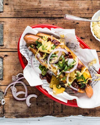 Laugen-Hotdog in einem roten Servierkörbchen mit Würstchen, Bacon, Zwiebeln und Ananas mit einem Schälchen Dijonnaise auf Holzuntergrund.