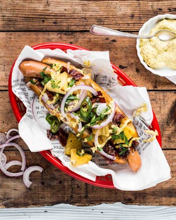 Laugen-Hotdog in einem roten Servierkörbchen mit Würstchen, Bacon, Zwiebeln und Ananas mit einem Schälchen Dijonnaise auf Holzuntergrund.