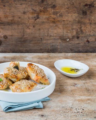 Griechische Kalitsounia mit Spinat-Füllung auf einem Teller neben Schälchen mit Öl und Salz auf einem Holztisch.