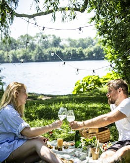 Zwei Menschen auf einer Picknickdecke am See, die mit Wein anstoßen.