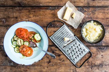 gehobelter Parmesan in einer Schüssel, daneben ein Stück Parmesan und eine Käsehobel. Links im Bild ein Teller mit Tomatenscheiben und gebratenen Zucchinischeiben