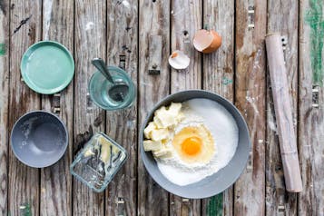 In einer grauen Schale Mehl mit Butter und einem Ei, drumherum leere Schalen und Behältnisse sowie Frischhaltefolie.