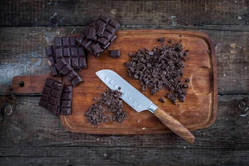 Gehackte Schokolade mit einem großen Küchenmesser auf einem Holzbrett. Links auf dem Brett liegt ungehackte Schokolade