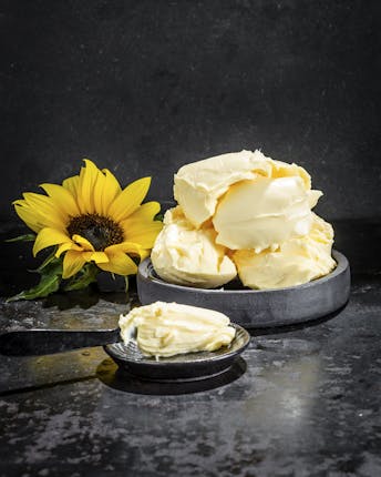 Ein geschmeidiger Berg von Margarine in einer dunklen Schale neben einem Messer mit Margarine und der gelben Blüte einer Sonnenblume vor schwarzem Untergrund