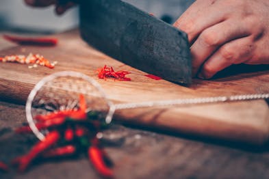 Chili wird auf einem Holzbrett mit einem großen dunklen Messer in feine Streifen geschnitten. Unscharf im Vordergrund einige Chilischoten