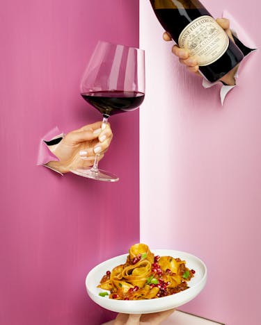 Drei Hände durchbrechen eine rosa Wand. Eine Hand hält eine Flasche, eine zweite ein Glas und eine dritte einen Teller mit Bandnudeln.