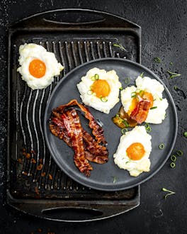 Cloud Eggs mit bacon auf einem schwarzen Teller. Dieser steht auf einem schwarzen Tablett.