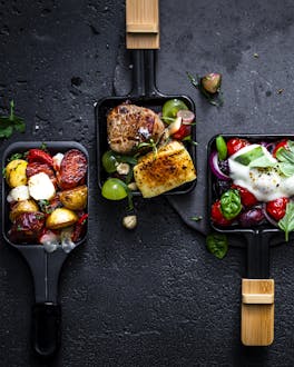 Vier bunt gefüllte Raclette-Pfännchen mit Gemüse, Fleisch und Käse