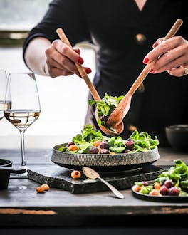 Zwei Hände richten mit Holz-Salatbesteck einen Feldsalat mit Weintrauben an. Daneben steht ein Teller mit Salat und zwei Gläser Weißwein.