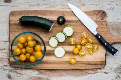 Eine Zucchini und einige Zucchini-Scheiben sowie gelbe Tomatenhälften auf einem Holzbrett mit einem Messer. In einer Glasschale gelbe Cherrytomaten