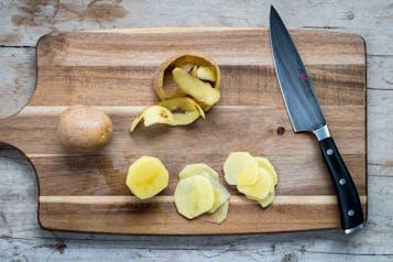 Eine teilweise geschälte Kartoffel liegt auf einem Holzbrett, daneben ein Messer