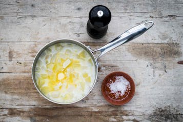 Sellerie- und Kartoffelwürfel mit geschmolzener Butter in einem Topf mit Stiel. Daneben eine schwarze Pfeffermühle und eine kleine rote Schale mit Salz.