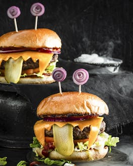Zwei Burger, die durch hängende Gurkenzungen und aufgerollten Zwiebelaugen aussehen wie Monster. Links im Bild eine rauchende Schüssel. Der Bildhintergrund ist schwarz