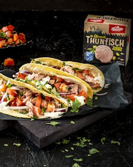Hintereinander liegen drei prall gefüllte Tacos mit Thunfisch, Pico de Gallo und Mayonnaise auf einem schwarzen Brett