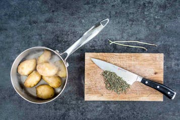 Im linken Bildabschnitt gekochte Kartoffeln in einem Topf mit Stiel. Im rechten Bildabschnitt ein Holzbrett mit gehacktem Rosmarin und einem großen Küchenmesser