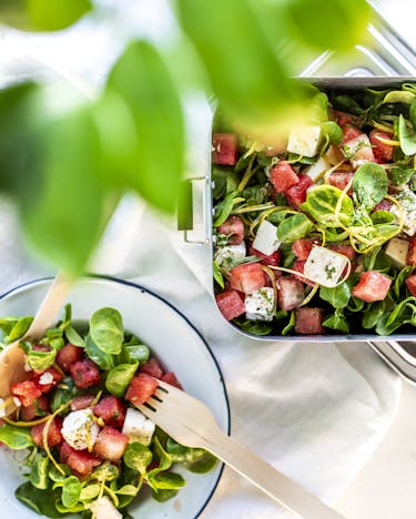 Lunchdose und weiße Schale mit Wassermelonen-Feta-Salat und Feldsalat.