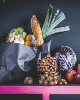 Eine dunkle Papiertüte mit Baguette und Gemüse neben offenen Metallkörben und einer kleineren Papiertüte mit weiteren frischen Lebensmitteln auf einem pink umrandeten Board vor einem schwarzen Hintergrund