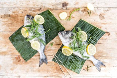 2 Fische liegen mit Dill und Zitronenscheiben auf Bananenblättern