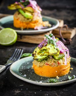 Süßkartoffel-Causa mit Quinoa und Avocado, das Schichtgericht ist angerichtet auf zwei farbigen Tellern und ein Teller ist auf einem Holzbrett drapiert, daneben eine halbe Limette, rote Zwiebeln und eine Gabel, fotografiert auf dunklem Untergrund.