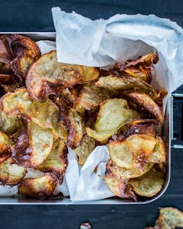 Knusprige, goldbraune, selbstgemachte Kartoffelchips auf Butterbrotpapier in glänzender Lunchbox, von oben fotografiert.