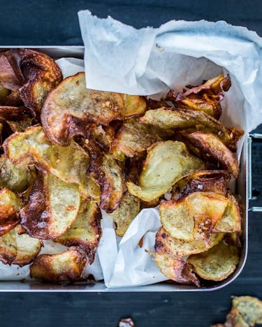 Knusprige, goldbraune, selbstgemachte Kartoffelchips auf Butterbrotpapier in glänzender Lunchbox, von oben fotografiert.