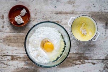 Ein aufgeschlagenes Ei in Mehl in einer großen Glasschale, daneben ein kleiner Topf mit Milch, geschmolzener Butter und Hefe.