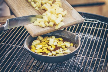 Kartoffelwürfel werden in einer großen Pfanne auf einem Grill gegart. Von einem Holzbrett werden Kartoffelwürfel mit einem Messerrücken in die Pfanne geschoben