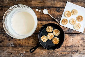 Schüssel mit Pancaketeig, fünf Mini Pancakes in einer Pfanne, daneben 6 Pancakes auf einem Küchenpapier auf Holzuntergrund