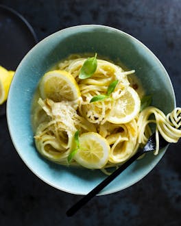 Eine türkise Schale mit Zitronen-Spaghetti, Zitronenscheiben und frischem Basilikum vor dunklem Untergrund