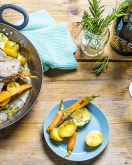 In Frühlingsfarben sind die Lammkeulen-Scheiben in einer Pfanne an Kartoffeln, Möhren und Zucchini auf einem hellen Holztisch mit eine türkisen Geschirrtuch präsentiert. Dabei steht ein Glas Prosecco.