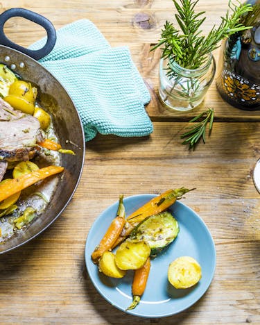 In Frühlingsfarben sind die Lammkeulen-Scheiben in einer Pfanne an Kartoffeln, Möhren und Zucchini auf einem hellen Holztisch mit eine türkisen Geschirrtuch präsentiert. Dabei steht ein Glas Prosecco.