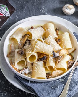 Paccheri mit Parmesansauce und Pilzen in weißem Teller auf dunklem Untergrund, im Hintergrund Parmesan und Champignons