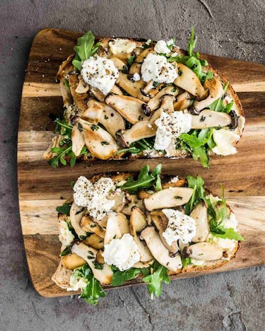 Zwei belegte Pilz-Stullen mit Salat auf einem Holzbrettchen auf grauem Untergrund.