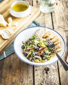 Weiße Schale mit Nudelsalat mit Tomaten, Rucola, Oliven und Parmesanhobeln neben geschnittener Hähnchenbrust auf Holztisch