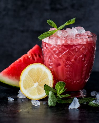 Glas mit Wassermelonen-Smoothie, Crushed Ice und Minze neben Stück Wassermelone und Zitronenhälfte auf dunklem Grund