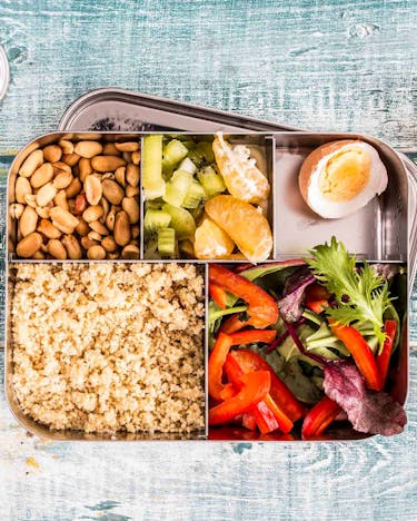 In einer Brotdose liegen Nüsse, Couscous, Obst, ein Ei und Salat. Auf Holz-Untergrund. Daneben ein Löffel
