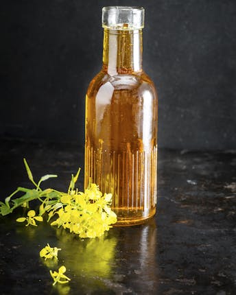 Eine kleine Glasflasche mit goldbraunem Rapsöl neben einer frischen gelben Rapsblüte vor dunklem Hintergrund