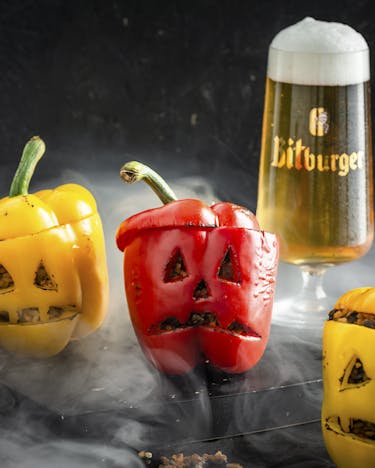 2 rote und 2 gelbe Paprikas mit eingeschnitzten Gesichtern, dahinter steht ein Glas Bier und darum herum Rauch