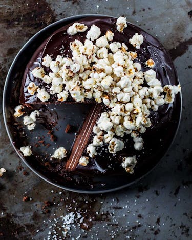 Angeschnittener Schokoladenkuchen mit Popcorn-Topping auf dunkler Servierplatte und Metalluntergrund.