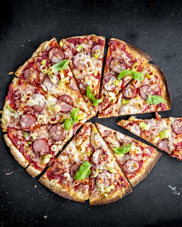 Eine runde Pizza mit krossem Rand und Belag aus Tomatensauce, Cabanossi, Frühlingszwiebeln und Käse liegt geschnitten auf schwarzem Untergrund.