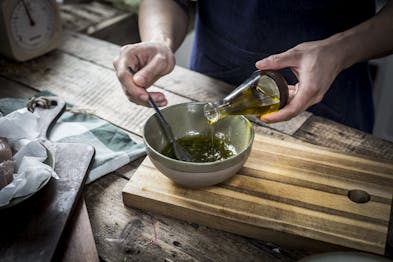 Aus einer Glasflasche wird Olivenöl untergerührt. Die Schale steht auf einem Holzbrett