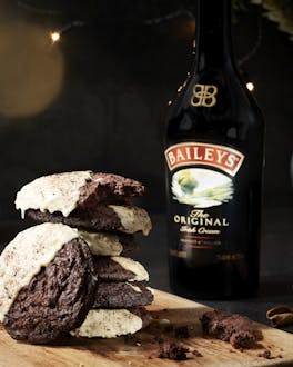 Ein Stapel Schokocookies auf einem Holzbrett. Eine Flasche Baileys steht im Hintergrund.
